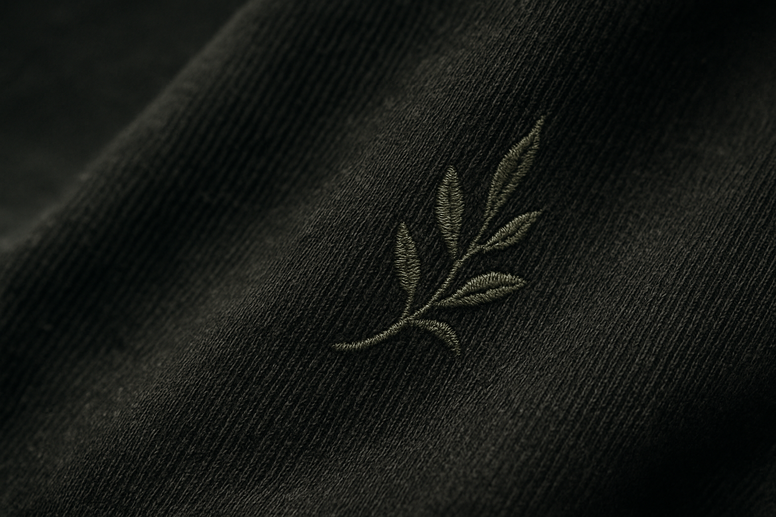 Close-up of dark grey woven fabric with a subtly embroidered olive branch, symbolizing hope and resistance, captured in soft natural light.